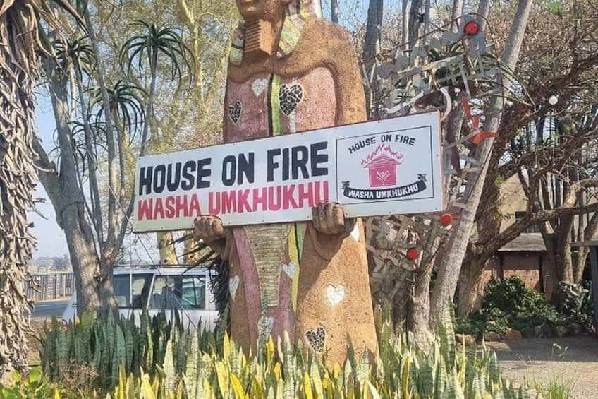 A large statue holding a sign that reads 'HOUSE ON FIRE WASHA UMKHUKHU.' 1