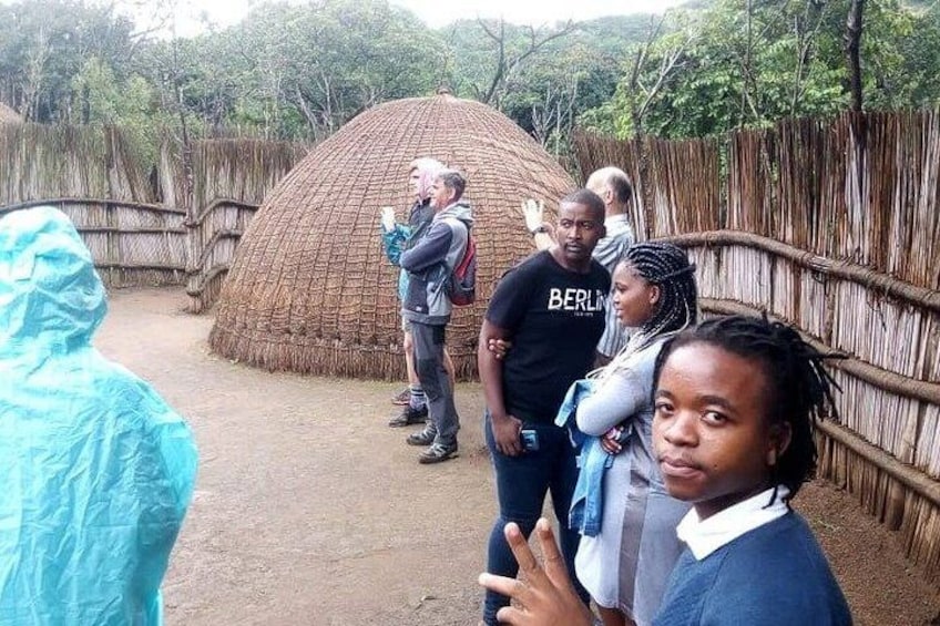 A group of people standing near a large, dome-shaped structure made of natural materials, possibly a traditional hut or exhibit, in an outdoor setting with a wooden fence and trees in the background. 5