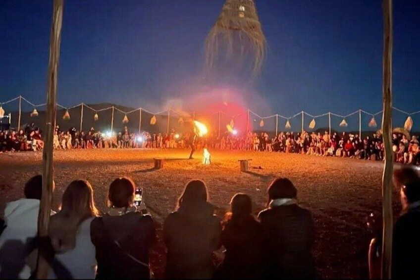 A nighttime outdoor event with a crowd gathered around a central fire, tents, and a large structure in the background. 4