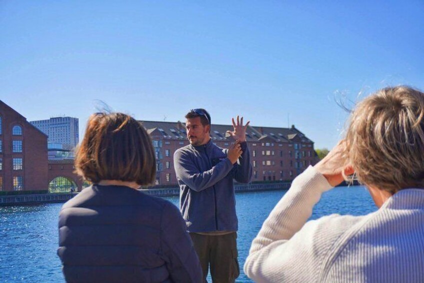 Three people on a boat, one gesturing, with a red brick building and water in the background. 1