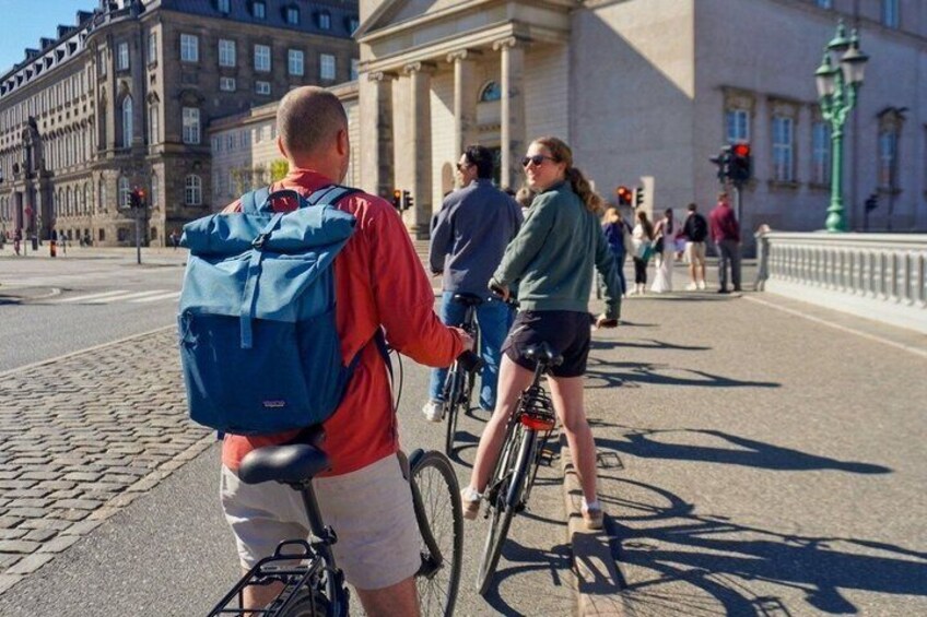 A man on a bicycle with a blue backpack, a woman on a bicycle, and a historic building in the background. 4