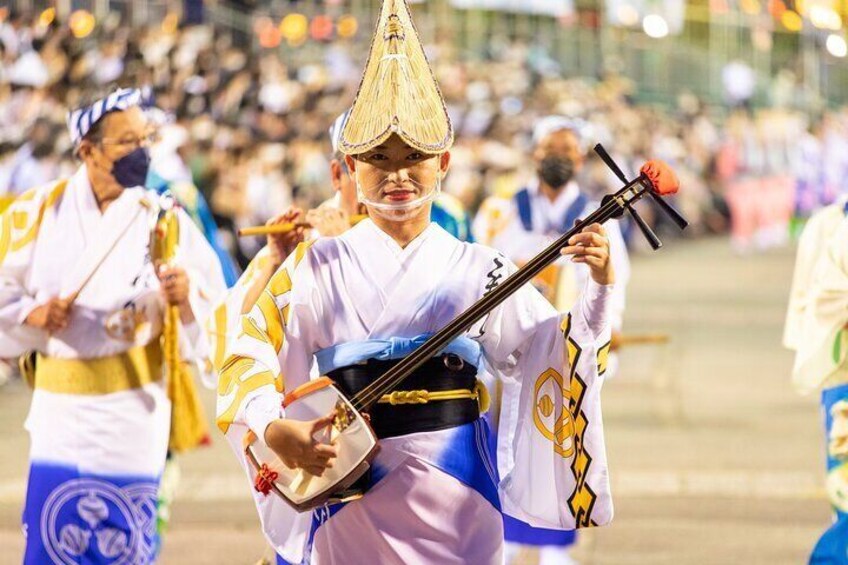 A person in traditional attire playing a stringed instrument, with a crowd in the background. 4