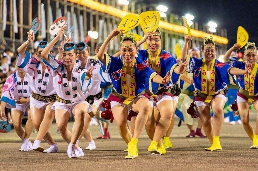 A group of people in colorful traditional attire, some holding fans, performing a synchronized dance on a sports field. 2