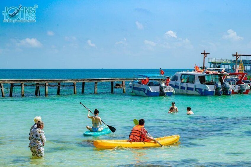 People kayaking and swimming in clear turquoise waters with a pier and boats in the background. 3