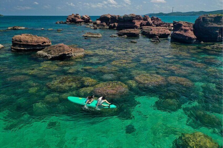 A person with a surfboard near clear turquoise water with rocks. 5