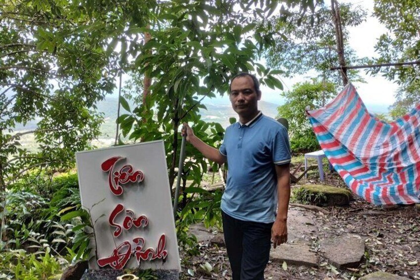 A man standing next to a sign with red lettering, a hammock, and a view of a landscape. 3