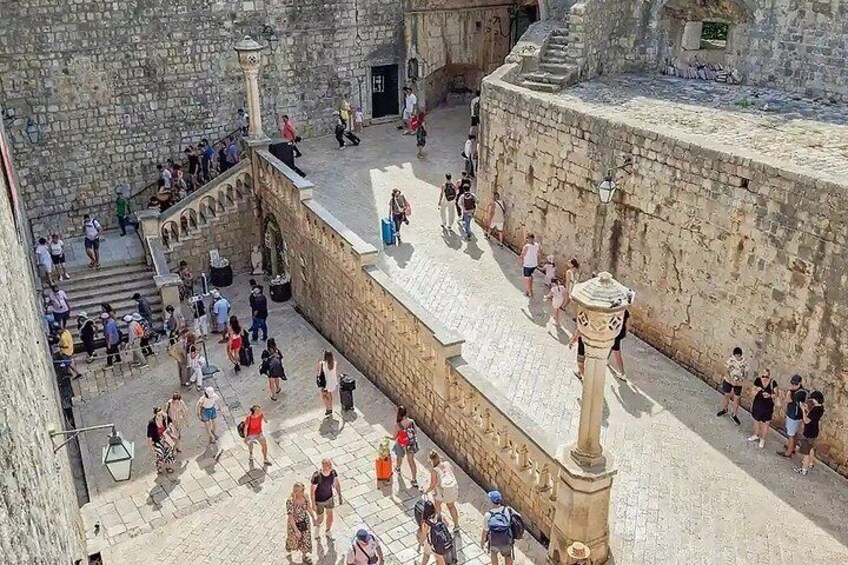 A crowded historical square with stone buildings, a central staircase, and a fountain. 3