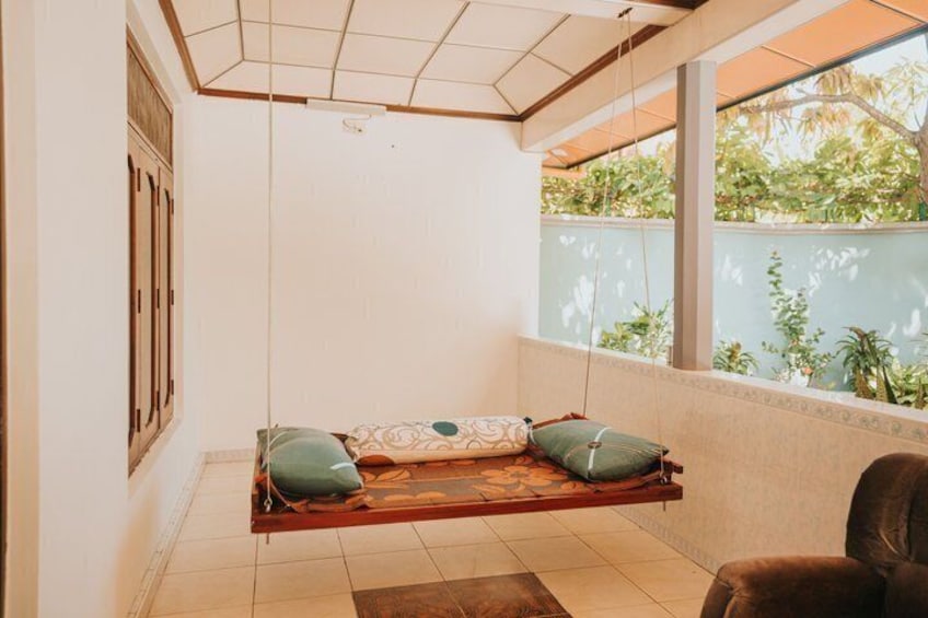 A room with a swing bed, a window with a view of greenery, and a tiled floor. 1