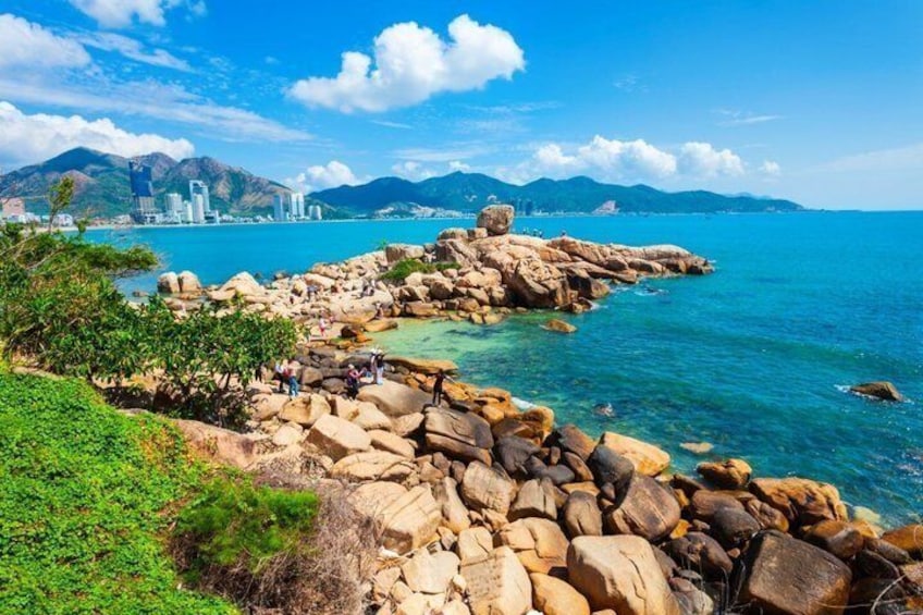 A coastal landscape with large rocks, clear blue water, and distant city buildings. 2