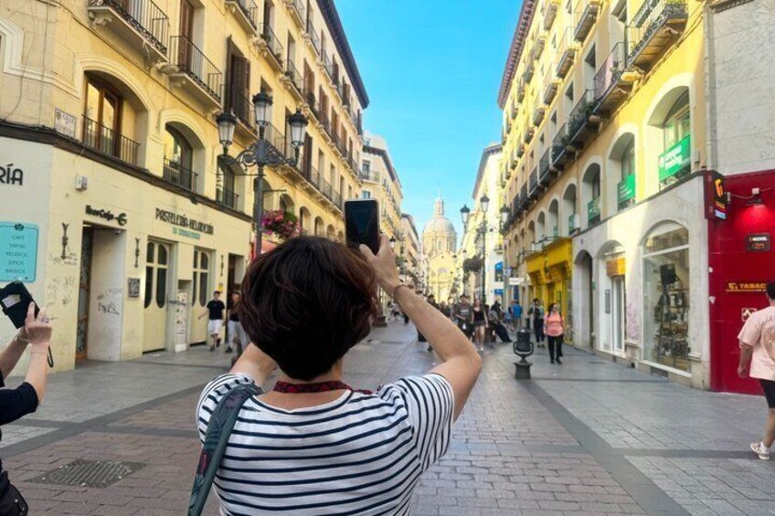 A person taking a photo with a smartphone in a pedestrian street lined with shops and buildings. 4