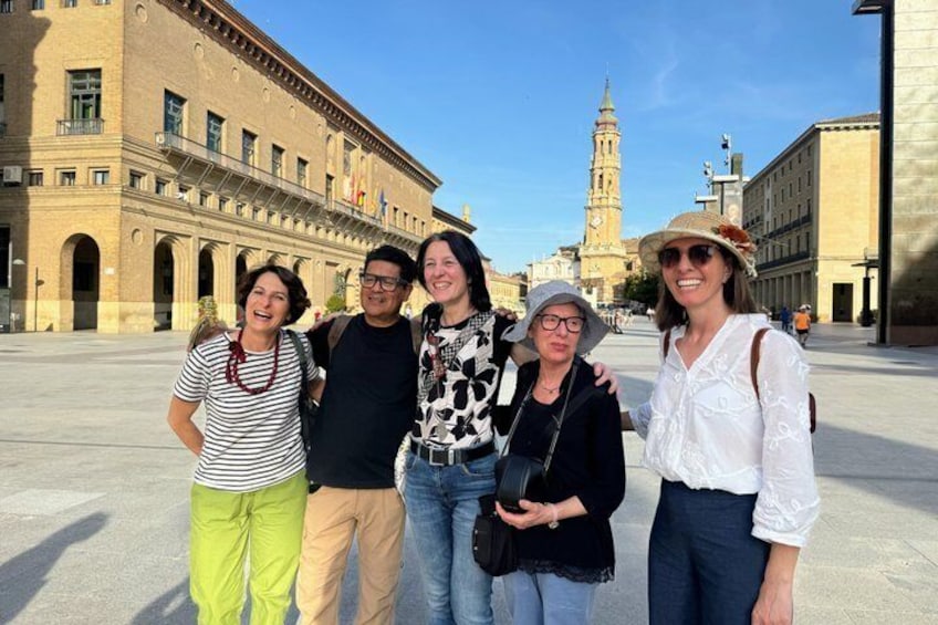 A group of people standing in a plaza with historical buildings and a clock tower in the background. 1