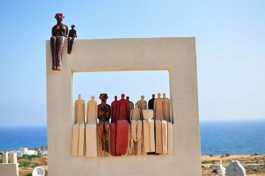 A sculpture of figures in various poses, a clear blue sky, and a distant view of the sea. 2