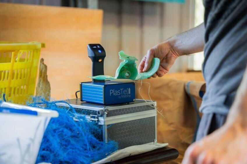 Iemand gebruikt een handapparaat op een blauwe doos met de tekst 'PlasTelf', met een gele mand en blauw materiaal op tafel. 4