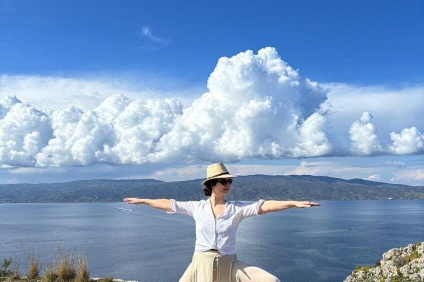 A person standing with arms outstretched on a cliff overlooking a body of water with mountains in the distance. 1