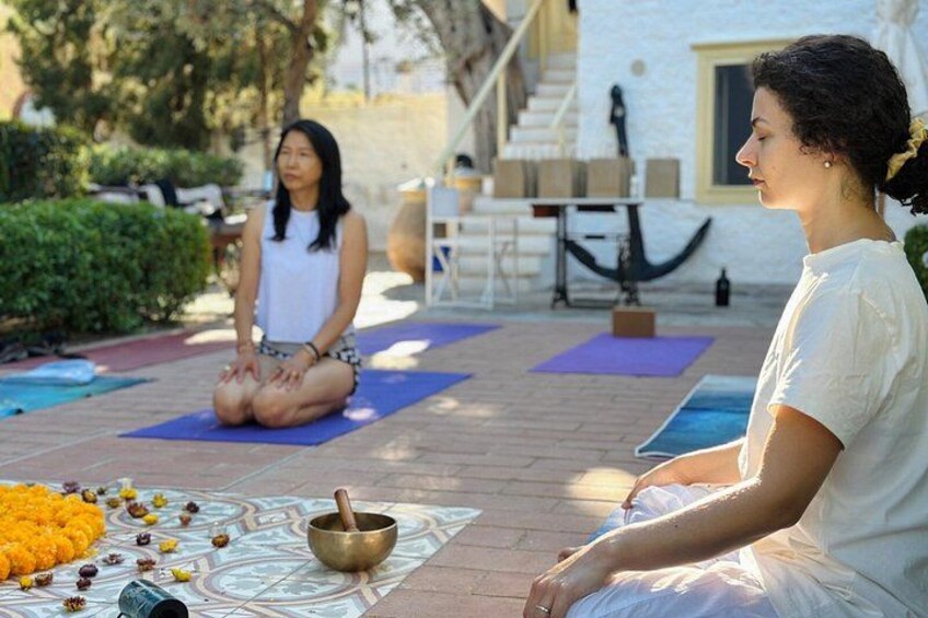 Two people sitting on yoga mats outdoors, with a decorative bowl and flowers on the ground. 3
