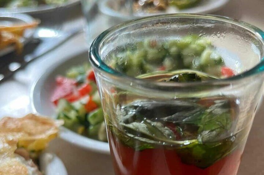 A glass of tomato juice with a green garnish, possibly herbs, and a blurred background with other dishes. 5