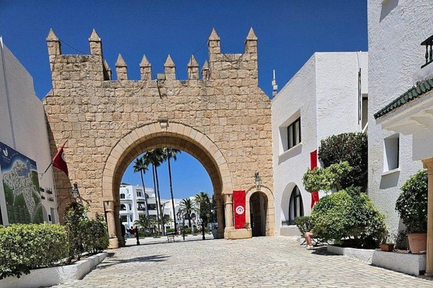 A stone archway with a flag, surrounded by buildings and palm trees. 1