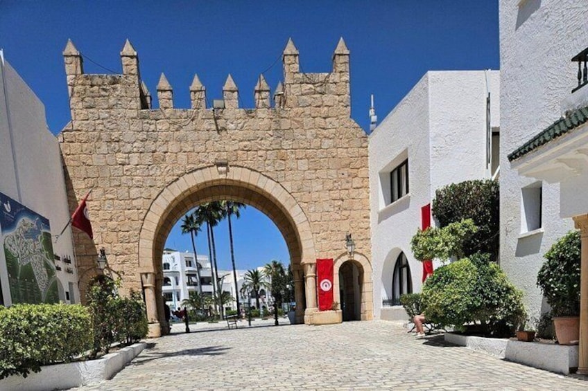 Un arco de piedra con una bandera roja, rodeado de edificios blancos y vegetación. 1