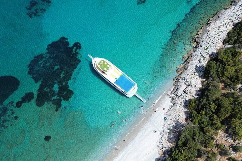 Vista aérea de un barco anclado en una playa con agua turquesa cristalina, una costa rocosa y una pequeña isla con vegetación. 1