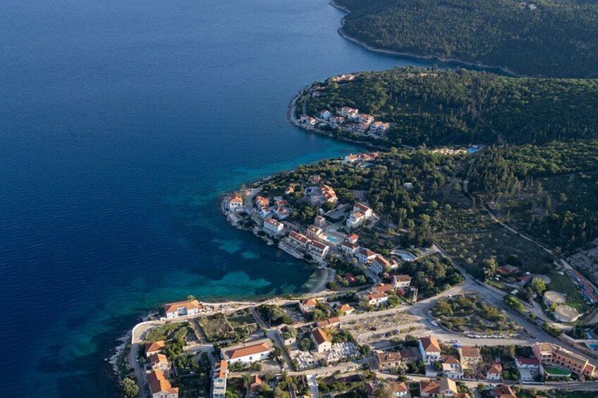 An aerial view of a coastal town with clear blue waters, a densely built area with red-roofed buildings, and a hilly landscape in the background. 3