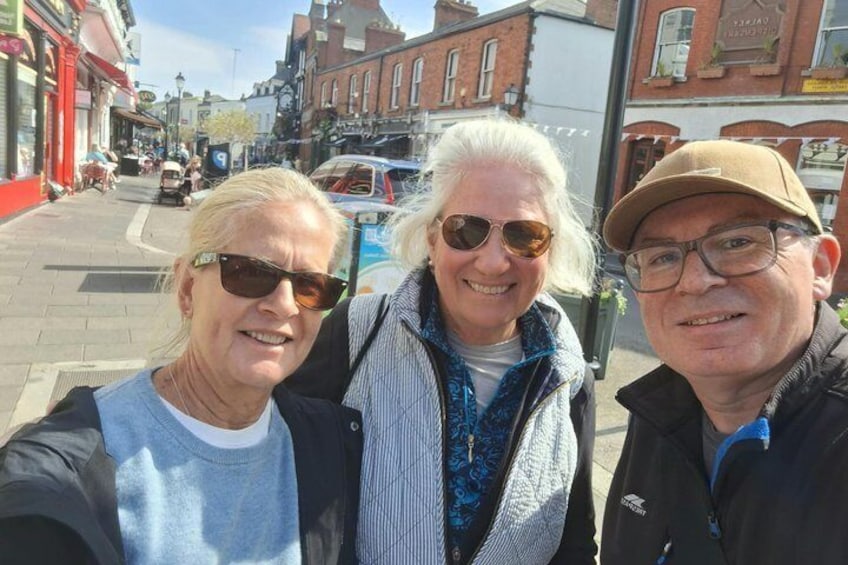 Three people taking a selfie on a city street with buildings and parked cars in the background. 2