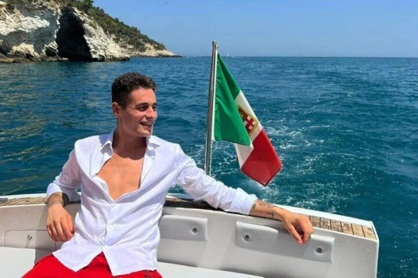 A person on a boat with a flag, water, and a rocky coastline. 2