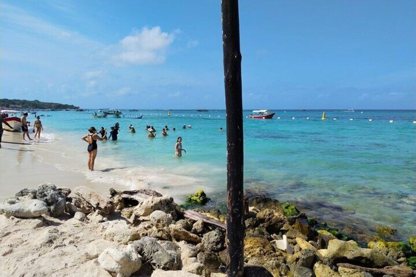 A beach with clear turquoise water, people swimming, and boats in the distance. 1