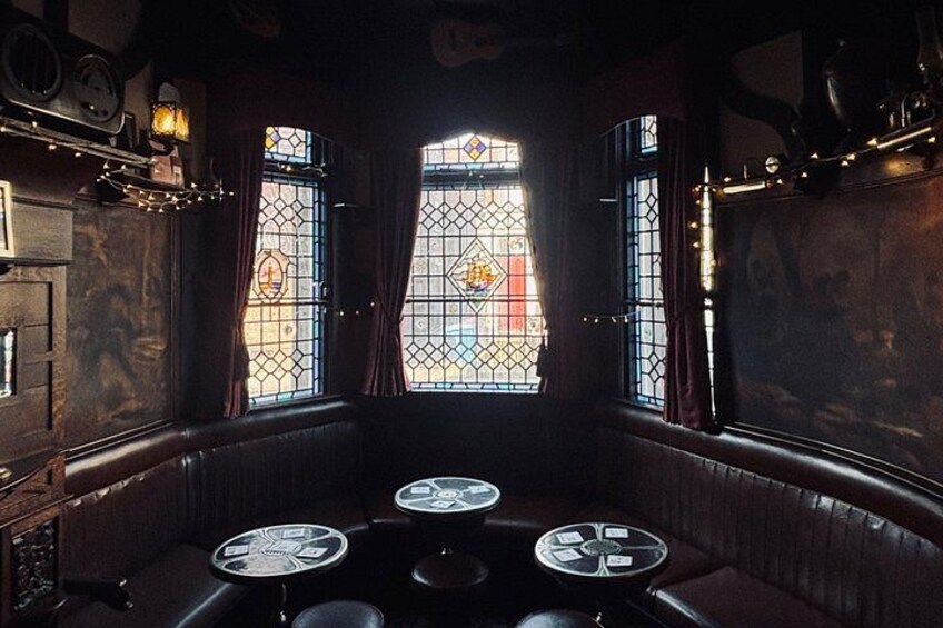 A dimly lit room with wooden walls, a central stained glass window, and a seating area with round tables. 2