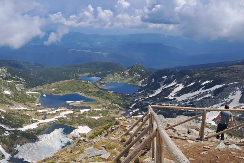 A mountainous landscape with a wooden viewing platform, a person standing, and a lake in the distance. 2