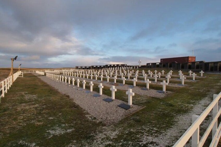 A cemetery with numerous white crosses arranged in rows, a fence, and a building in the background. 1