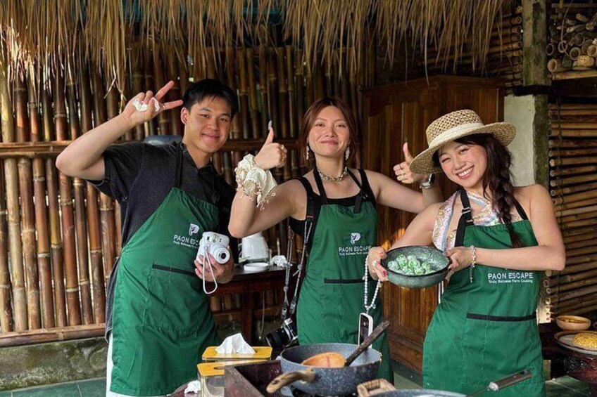 Three people in green aprons with 'PAON ESCAPE' branding, cooking outdoors with bamboo structures in the background. 2