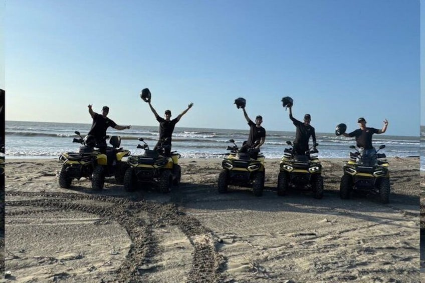 Vier personen op quad bikes op een zanderig strand, met de oceaan op de achtergrond. 4