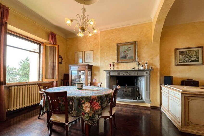 A dining room with a round table, chairs, a chandelier, a fireplace, and a window with curtains. 3