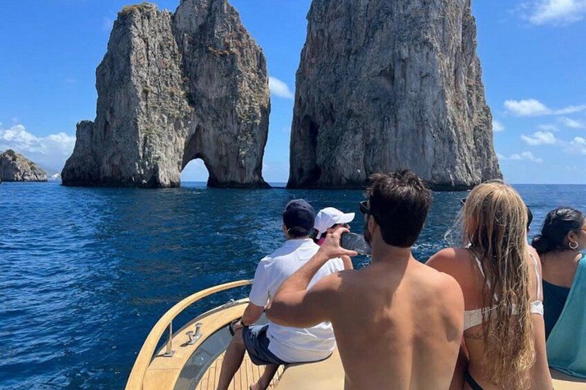 A boat with people on a clear day, a large rock formation with a natural arch, and calm blue water. 5