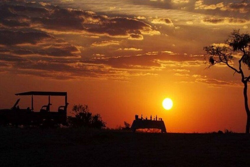 A sunset over a savanna landscape with silhouettes of a vehicle and a building. 1