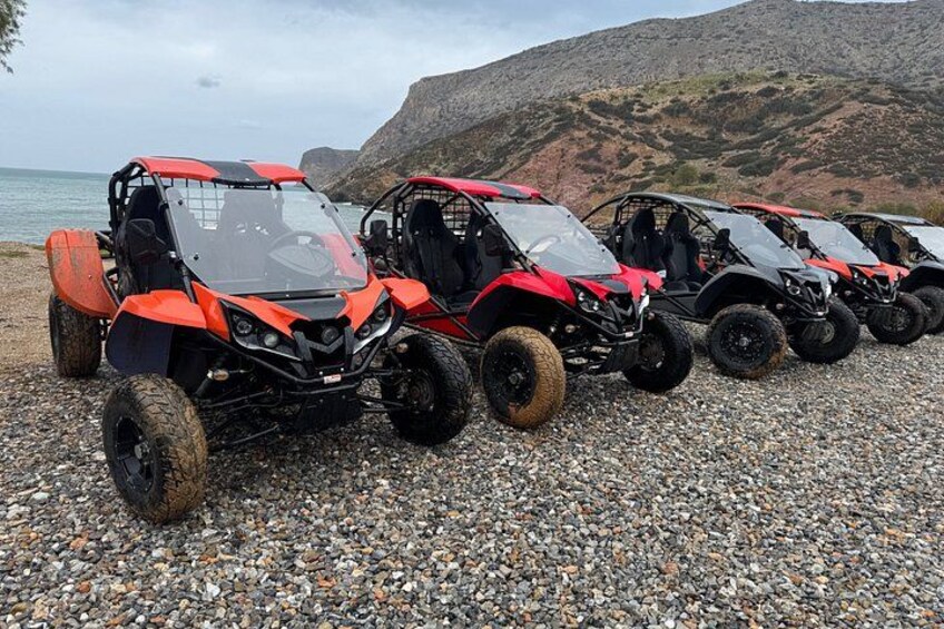 A row of red and black off-road vehicles parked on a pebbled beach with a mountainous backdrop. 2