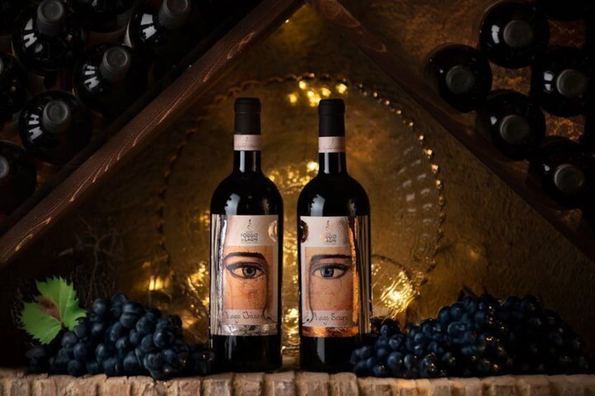 Two wine bottles with labels featuring an eye design, surrounded by grapes and a wooden wine rack. 1