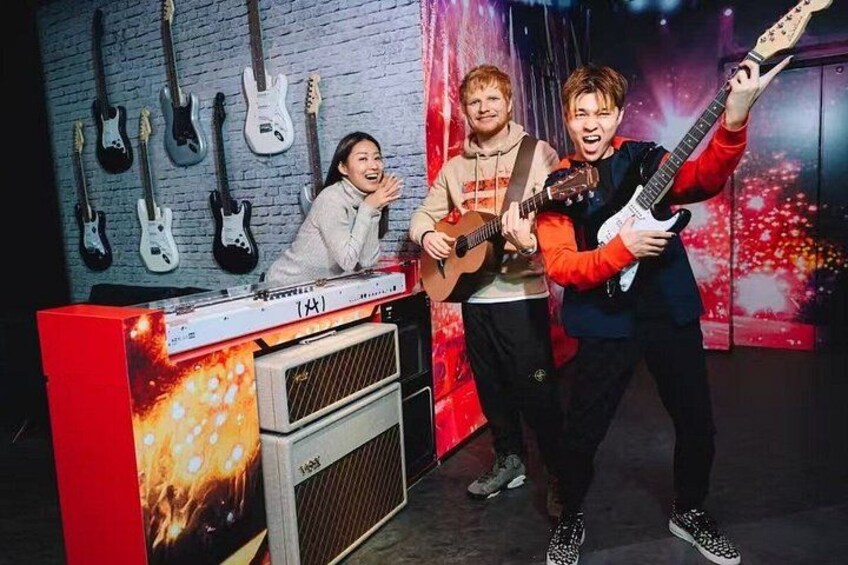 A person playing an electric guitar, another holding a guitar, and a third person standing behind a music stand with guitars on the wall. 4