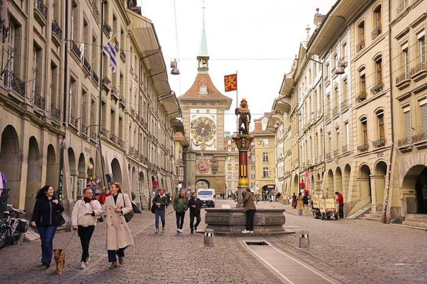 A cobblestone street lined with historic buildings, a central fountain, and a clock tower in the background. 1