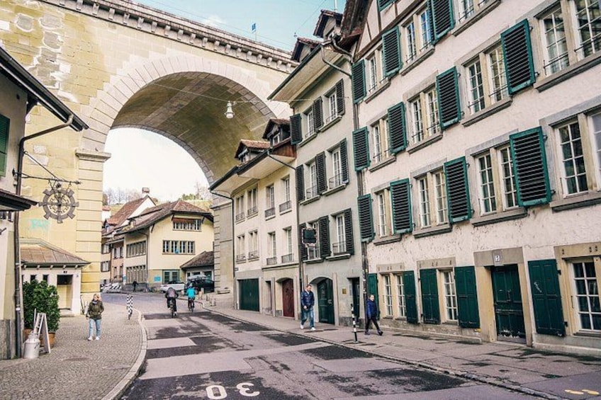 A cobblestone street with historic buildings, a stone arch bridge, and pedestrians. 2