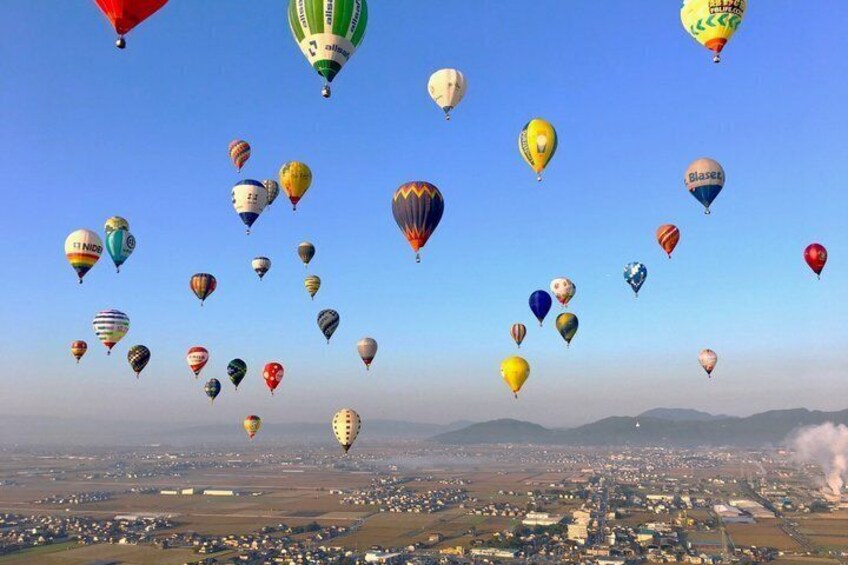 A variety of colorful hot air balloons over a landscape with buildings and smoke in the distance. 2