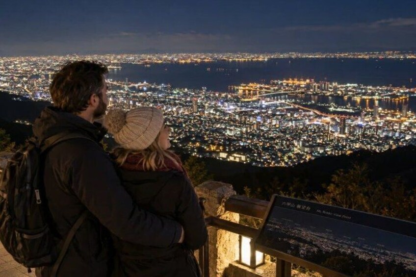 A couple overlooking a cityscape at night with illuminated buildings and a distant view of the sea. 4