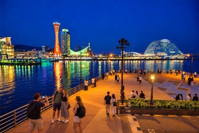 A waterfront promenade with people walking, a brightly lit tower, and a modern building with a dome. 5