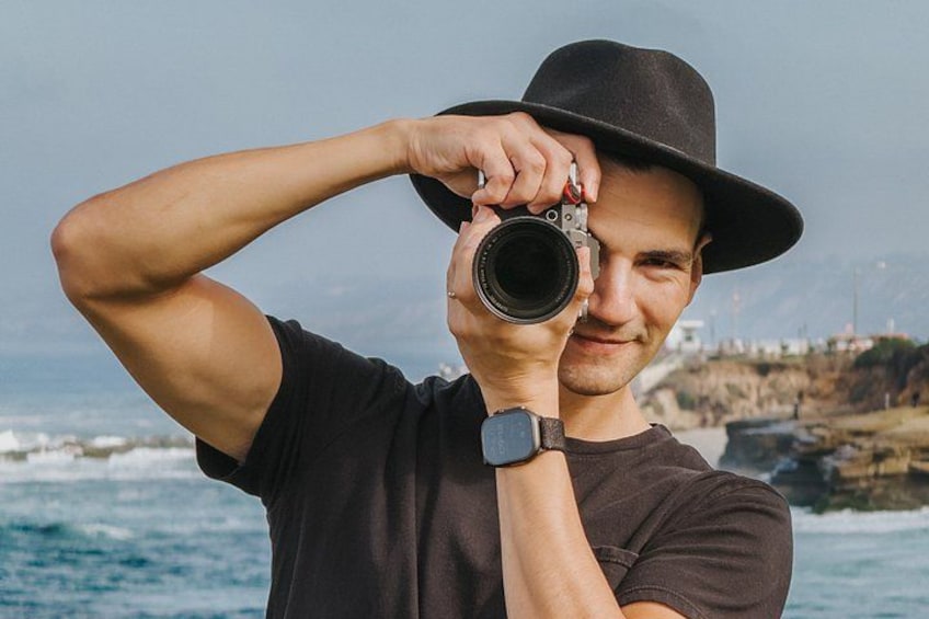 A person holding a camera with a black hat, wearing a dark shirt and a watch, standing by the ocean with rocky cliffs in the background. 1