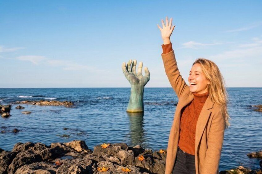 A person standing on rocky terrain by the sea, with a large hand sculpture in the water. 1