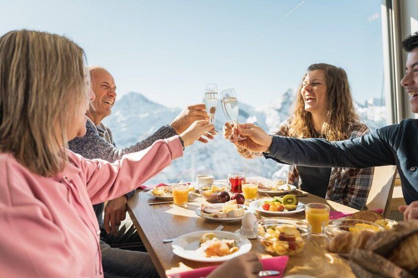 A group of people toasting with glasses at a table with breakfast food and a mountain view in the background. 1