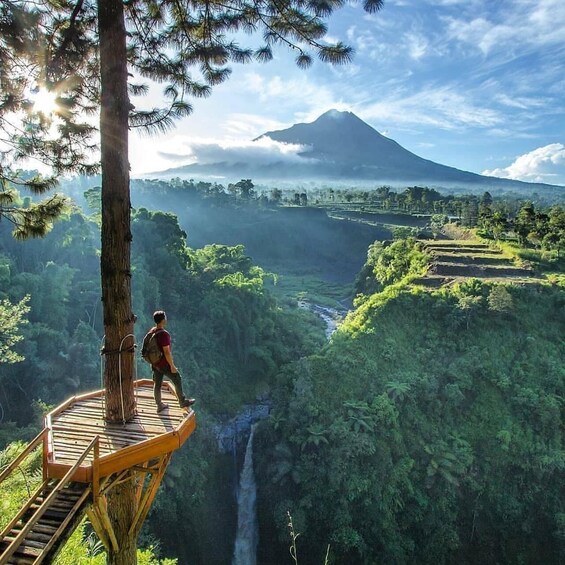 A person on a wooden platform overlooking a lush valley with a waterfall and a mountain in the background. 1