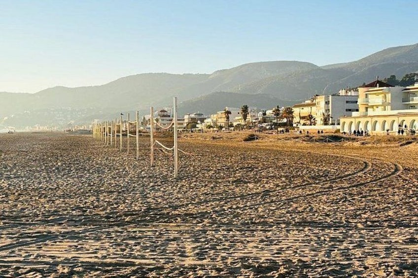 A pebbled beach with a fence, buildings, and mountains in the background. 2