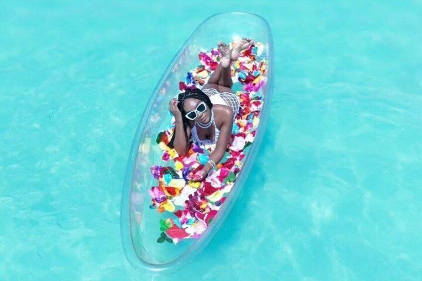 A person floating in a clear plastic raft filled with colorful flowers in a turquoise pool. 1