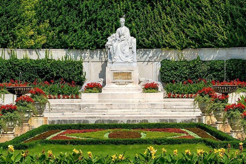 En statue på en sokkel med en mindetavle, omgivet af en velanlagt have med geometriske blomsterbede og en hæk i baggrunden. 3